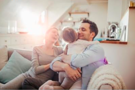 A joyful family sits on a couch. A young child hugs her father while her mother smiles happily beside them, bathed in sunlight.